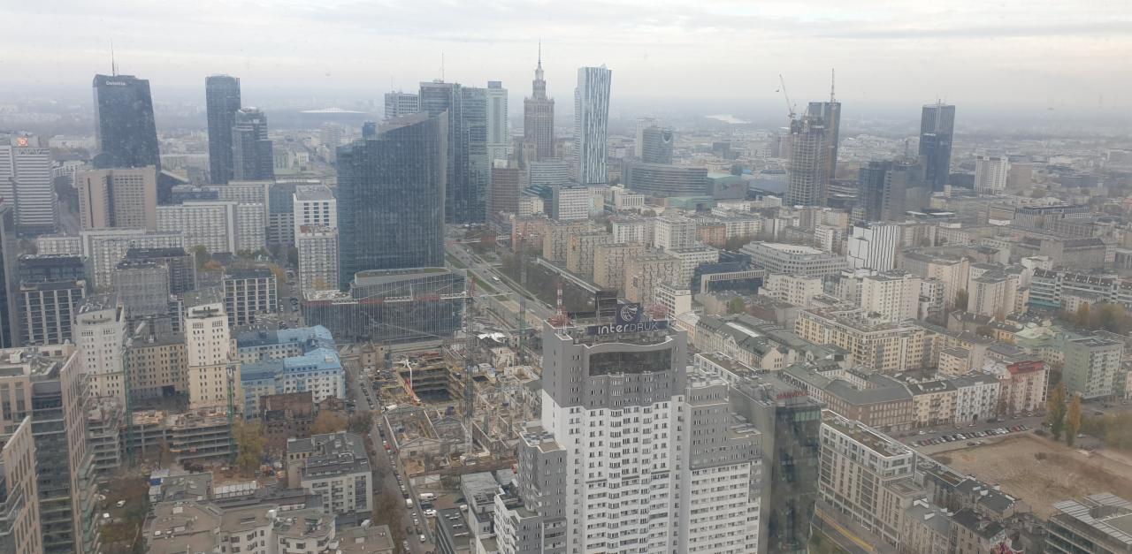 Panorama Warszawy z wysokości, widok na budynki w różnym stylu architektonicznym, w tym wieżowce, oraz place budowy w pochmurny dzień.