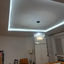 Salon z podwieszonym sufitem z oświetleniem LED, kryształowy żyrandol, pomarańczowa sofa i biała szafa.