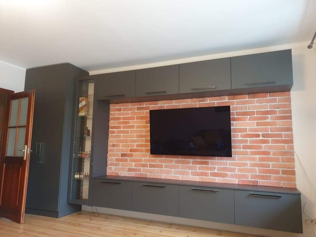 Szara zabudowa meblowa z szafkami wiszącymi i stojącymi na tle ściany z cegły, z telewizorem na środku, uzupełniona o przeszkloną witrynę z oświetleniem wewnętrznym i drewniane drzwi po lewej stronie.