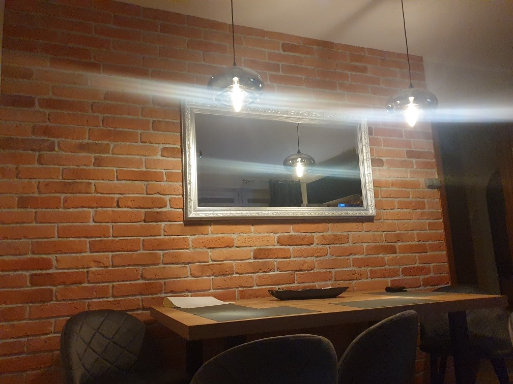 Ceglana ściana z lustrem w srebrnej ramie, oświetlona przez dwie szklane lampy wiszące, stół z szarymi krzesłami w tle, ciepłe światło.
