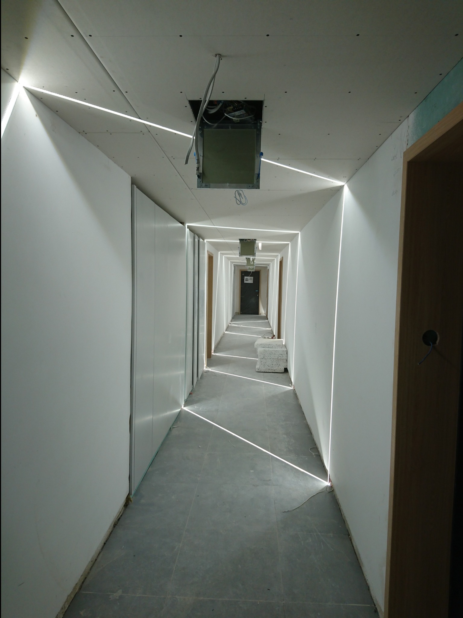 Długi korytarz w trakcie wykańczania, z zamontowanymi płytami gipsowo-kartonowymi na ścianach i suficie, oświetlony liniami LED wzdłuż krawędzi, widoczne niedokończone instalacje elektryczne...