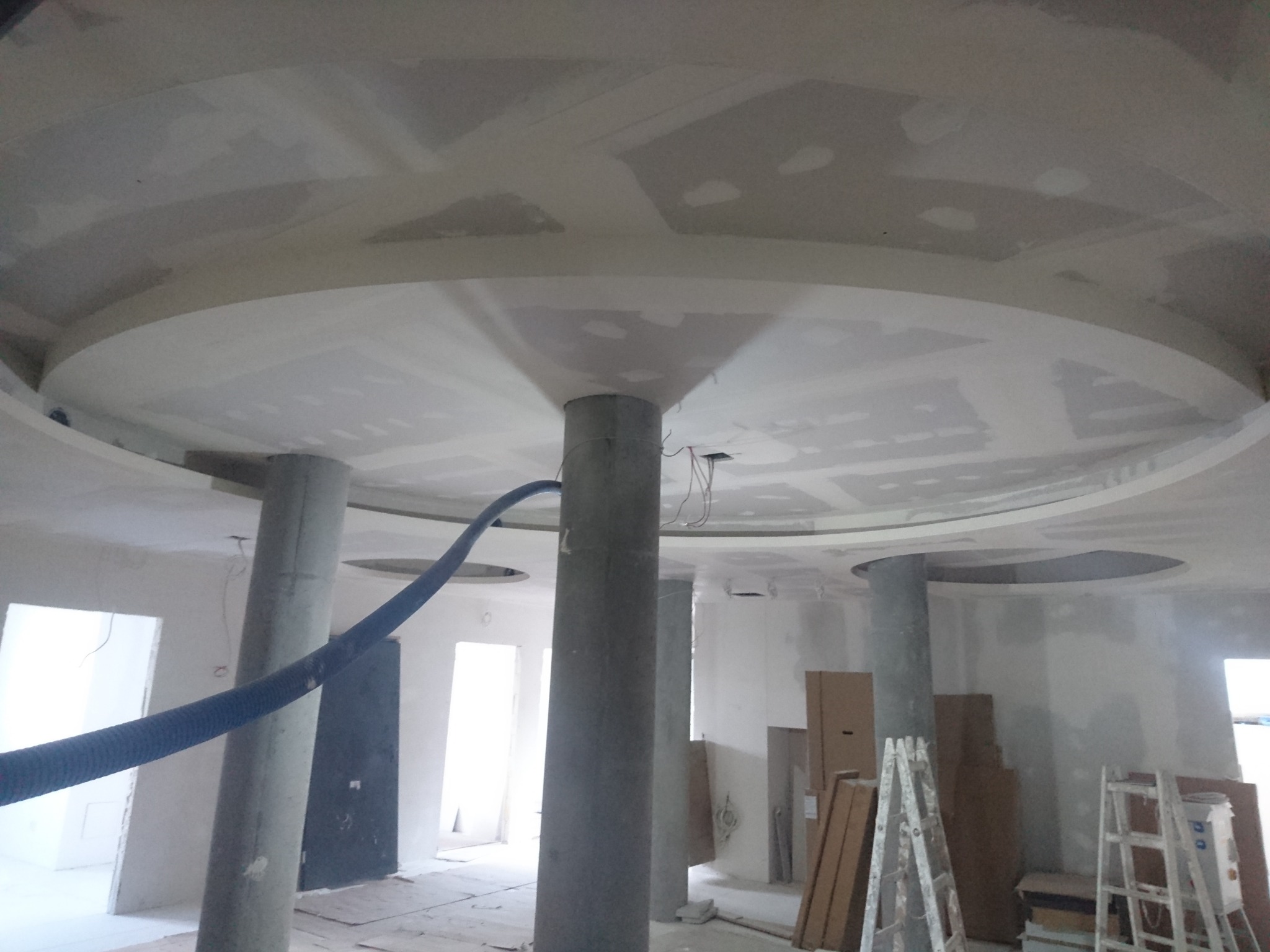 Sufit podwieszany z okrągłymi elementami i kolumnami w trakcie montażu, widoczne przewody i niebieski wąż wentylacyjny, surowy stan wykończenia.