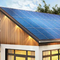 Nowoczesny dom jednorodzinny z panelami słonecznymi na dachu, oświetlony ciepłym światłem, z widocznym kominem i sąsiednim budynkiem w tle.