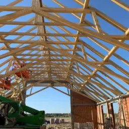 Wznoszenie drewnianej więźby dachowej z użyciem podnośników koszowych na tle błękitnego nieba i częściowo wymurowanej ściany budynku.