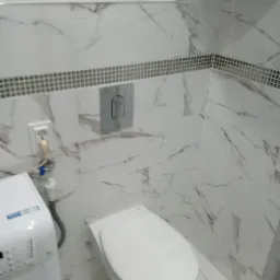 Wykończona łazienka z toaletą podwieszaną i pralką, ściany wyłożone białymi płytkami imitującymi marmur z dekoracyjnym pasem mozaiki.