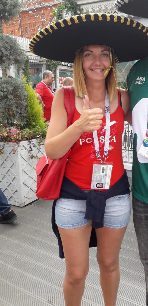 Uśmiechnięta kobieta w sombrero i koszulce z napisem POLSKA, z identyfikatorem na szyi, unosi kciuk w górę na tle czerwonej ceglanej architektury i zieleni.