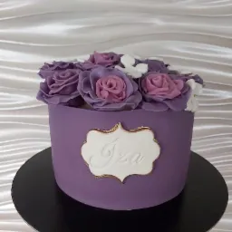 Fioletowy tort z lukrowanymi różami i napisem 'Iza' na białej etykiecie z obwódką w kolorze starego złota, umieszczony na czarnym podeście.