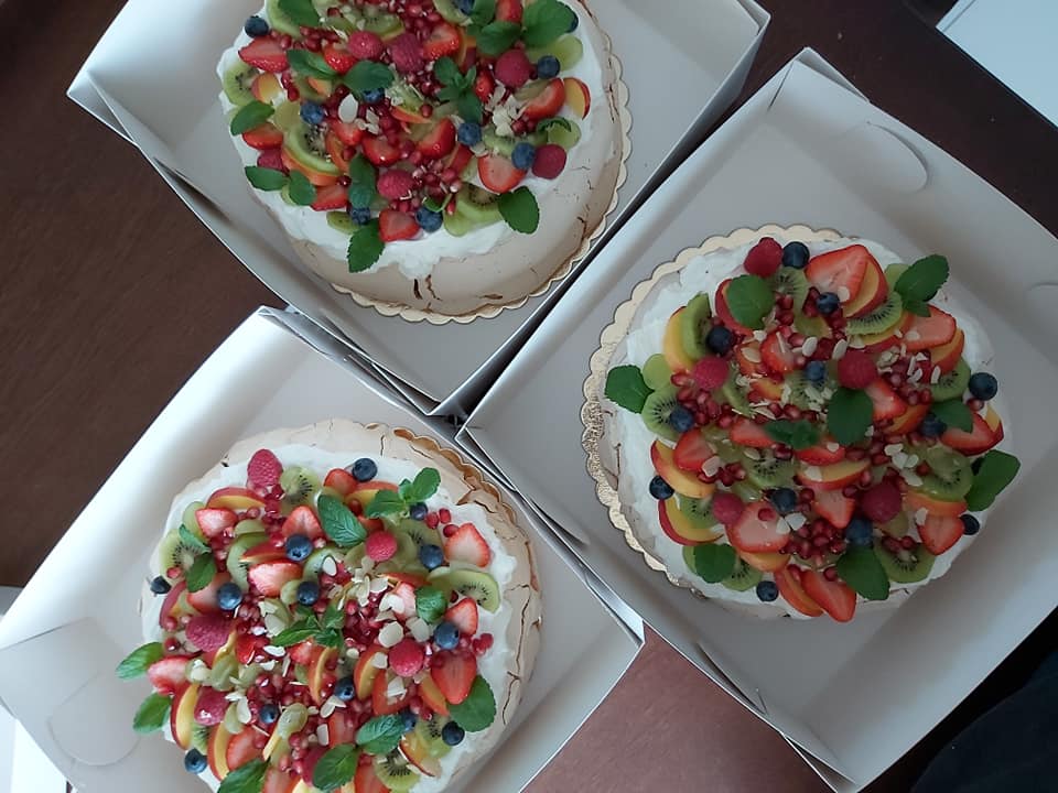 Trzy torty Pavlova w kartonowych pudełkach, udekorowane bitą śmietaną, truskawkami, kiwi, borówkami, malinami, granatem i listkami mięty.