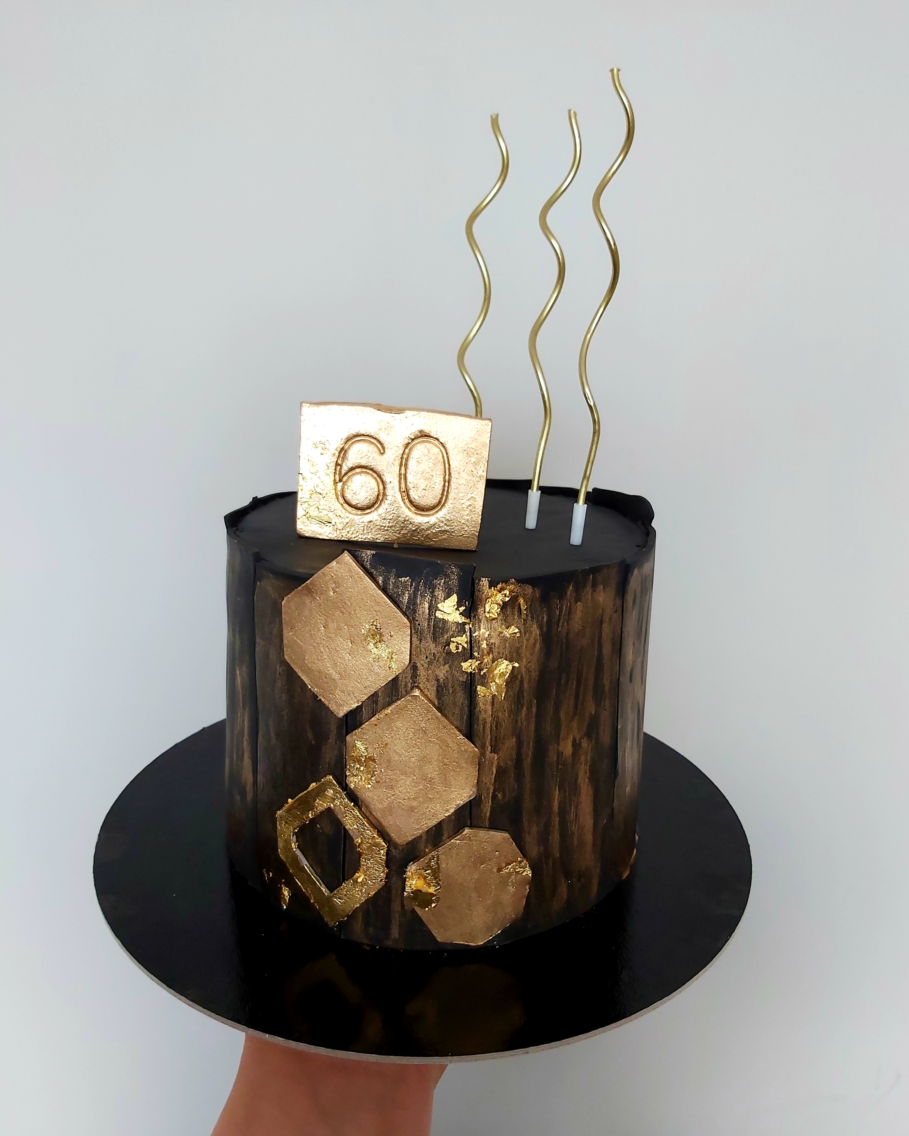 Tort urodzinowy w stylu rustykalnym z czarną polewą, złotymi dekoracjami geometrycznymi i napisem '60' na górze, umieszczony na czarnym talerzu.