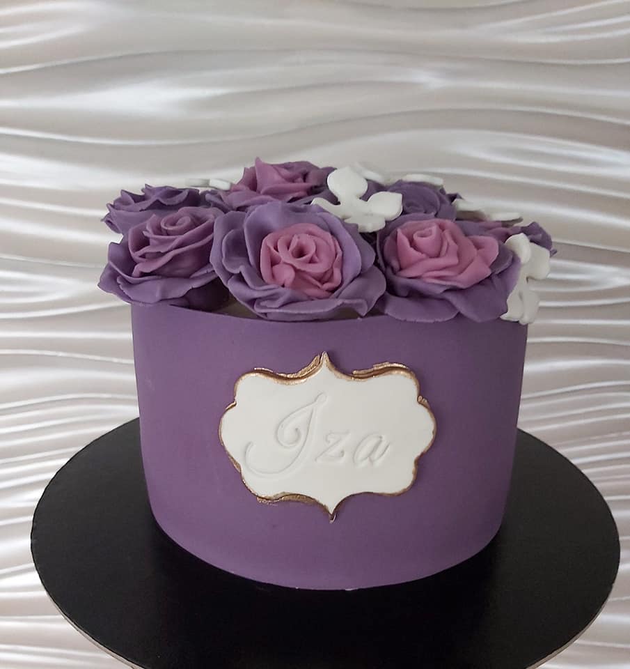 Fioletowy tort z lukrowanymi różami i napisem 'Iza' na białej etykiecie z obwódką w kolorze starego złota, umieszczony na czarnym podeście.