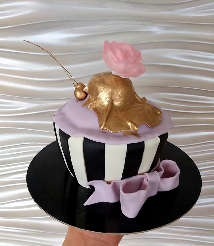 Dekoracyjny babeczka w czarno-białe paski z fioletowym lukrem, złotą polewą, różową różą z cukru i fioletową kokardą, prezentowana na czarnym talerzu.