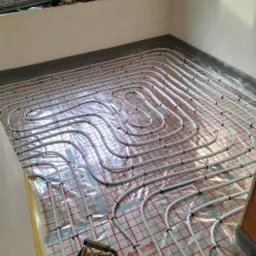 Białe rury ogrzewania podłogowego ułożone na folii z czerwoną siatką, widoczny zwijak z żółtym kablem i fragment okna w tle.