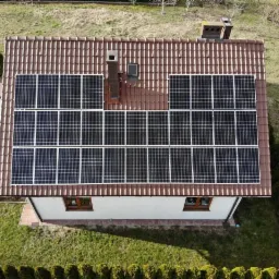 Widok z góry na dom z brązową dachówką, na której zamontowano panele fotowoltaiczne; dwa kominy na dachu.