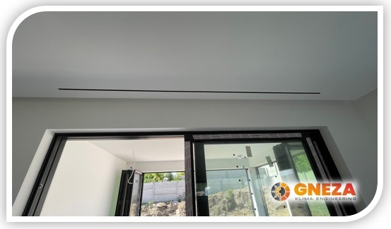 Dyskretny, liniowy nawiewnik sufitowy w nowoczesnym wnętrzu z dużym oknem przesuwnym, widoczne logo firmy instalacyjnej w prawym dolnym rogu okna.