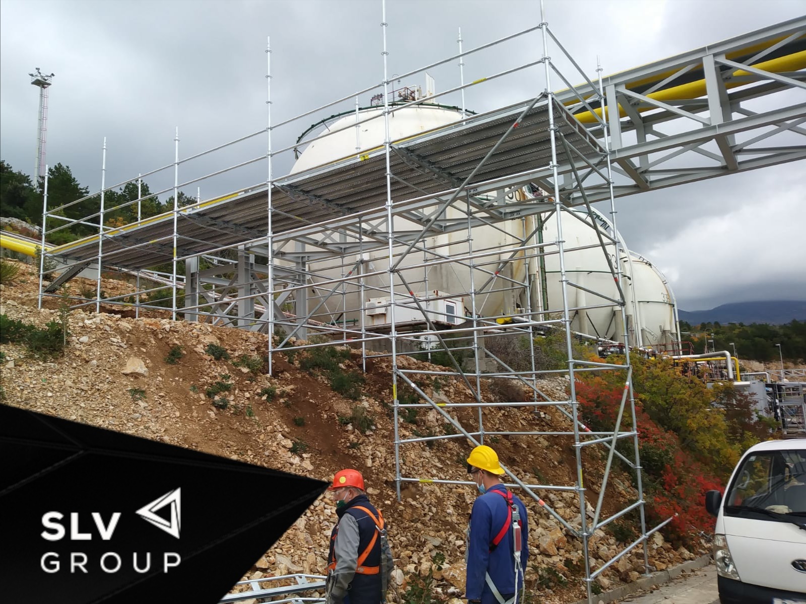 Montaż rusztowania przy zbiornikach przemysłowych, dwóch pracowników w kaskach i z zabezpieczeniami, logo firmy w lewym dolnym rogu.