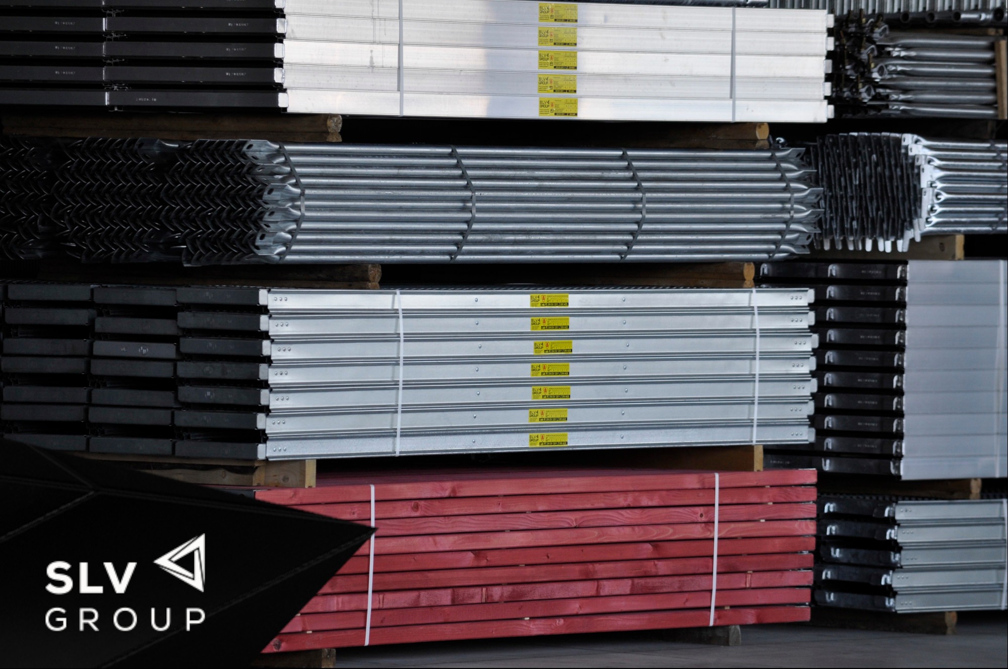 Stosy metalowych i drewnianych elementów konstrukcyjnych różnych typów i kolorów, zabezpieczonych na paletach, z widocznym logo SLV Group w lewym dolnym rogu.