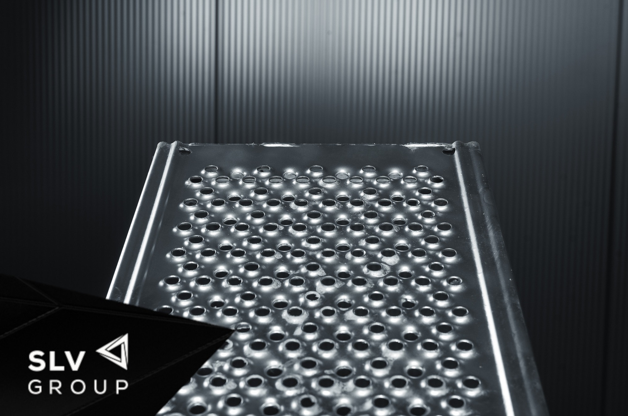 Metalowa platforma robocza z perforowaną powierzchnią, oświetlona chłodnym światłem, z logo SLV Group w lewym dolnym rogu, na tle pionowych, metalowych paneli.