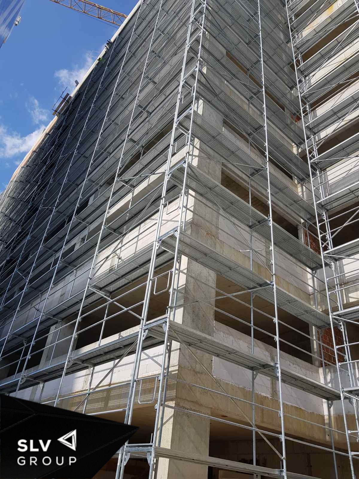Wysoki budynek w trakcie budowy, osłonięty rusztowaniami z siatką ochronną, widok pod kątem z dołu, logo firmy w lewym dolnym rogu.