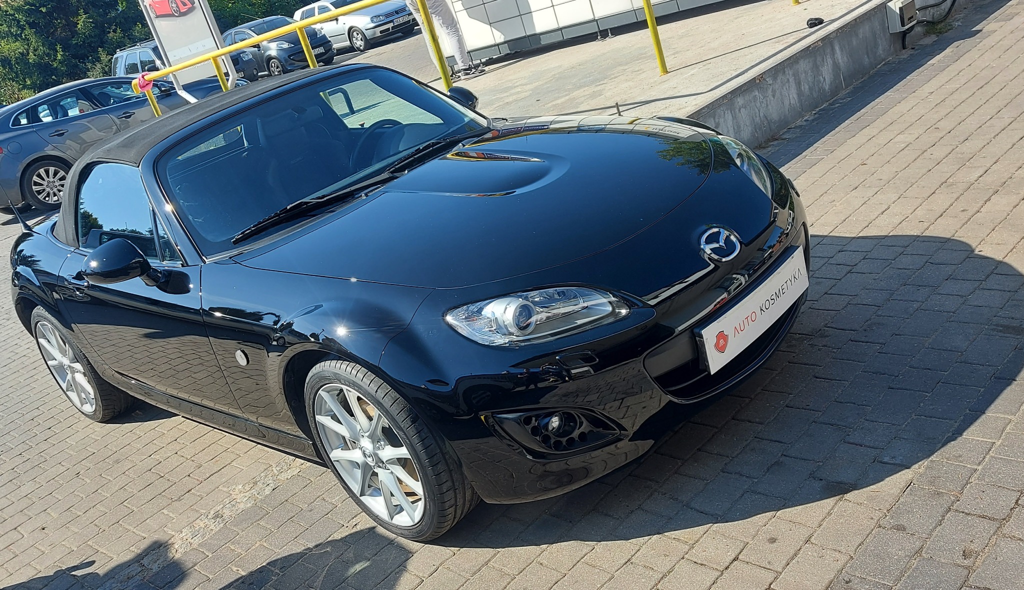 Błyszcząca, czarna Mazda MX-5 po umyciu, zaparkowana na kostce brukowej, z widocznym logo 'Auto Kosmetyka' na tablicy rejestracyjnej.