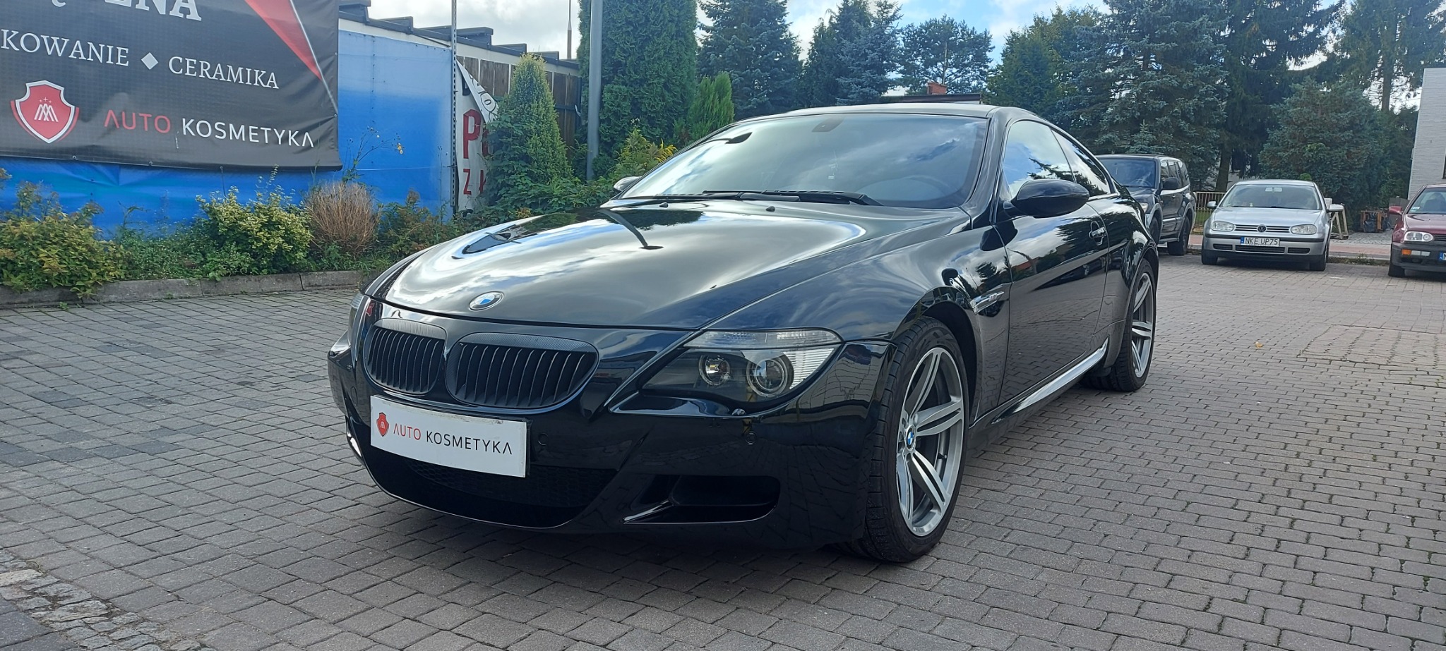 Czarna, lśniąca BMW serii 6 po zabiegach auto kosmetyki, zaparkowana na brukowanym placu, z widocznym banerem reklamowym firmy w tle.