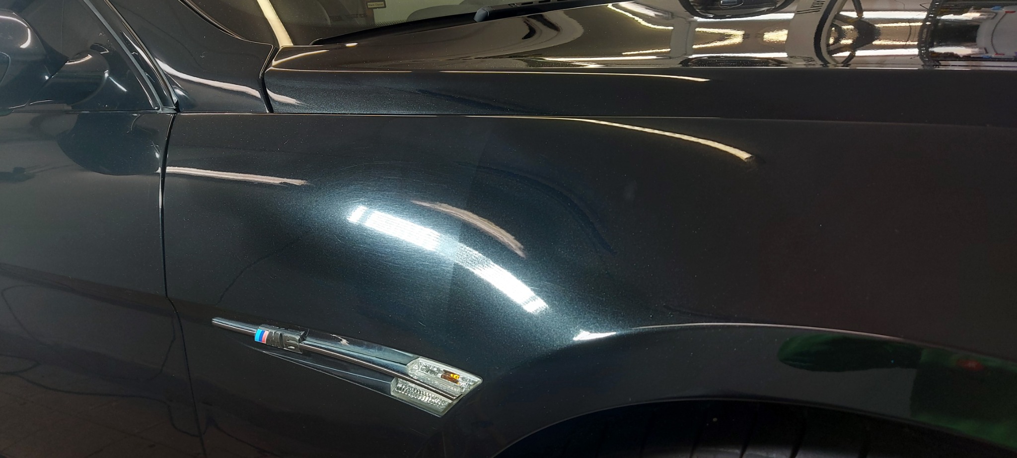 Fragment karoserii ciemnego samochodu z widocznymi śladami polerowania i hologramami, reflektory boczne włączone.