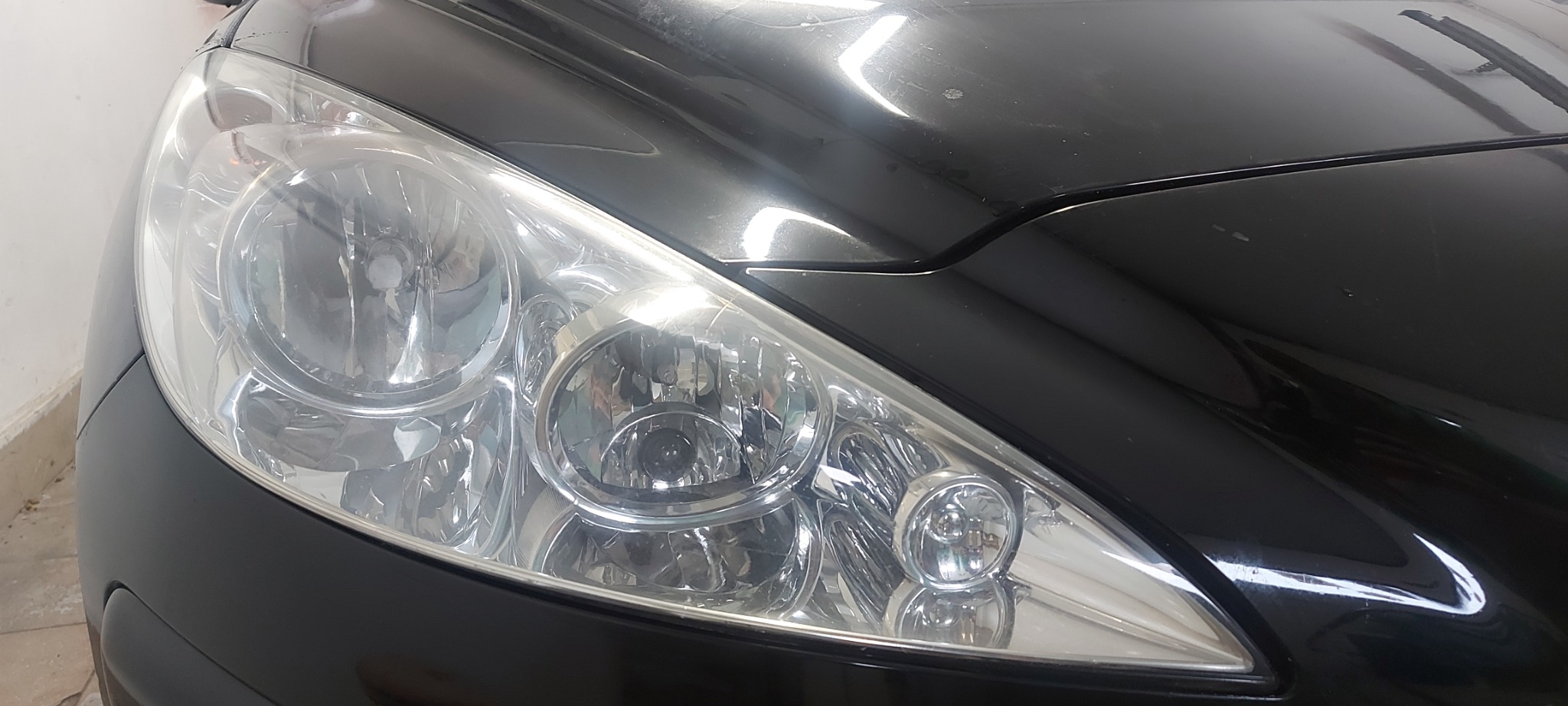 Błyszczący reflektor czarnego samochodu, widoczne drobne rysy na karoserii, światło odbija się od powierzchni.