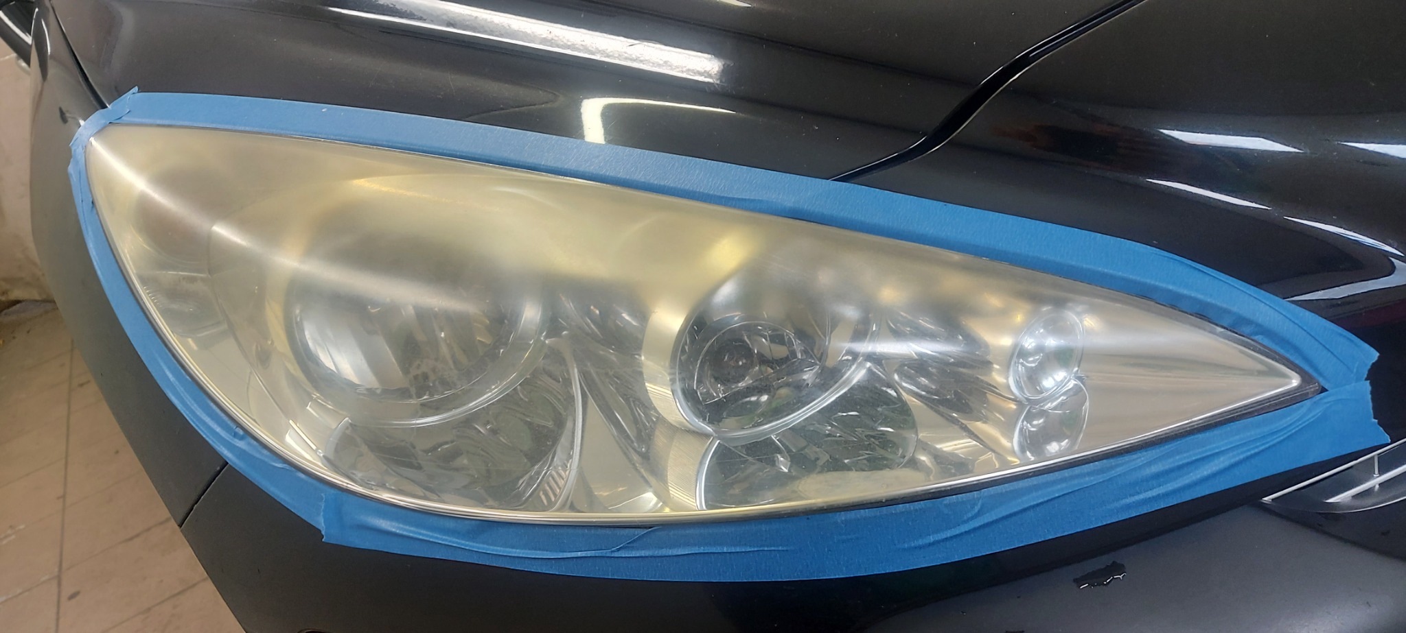 Żółta, zmatowiała lampa samochodowa zabezpieczona niebieską taśmą malarską przed polerowaniem na czarnym samochodzie.