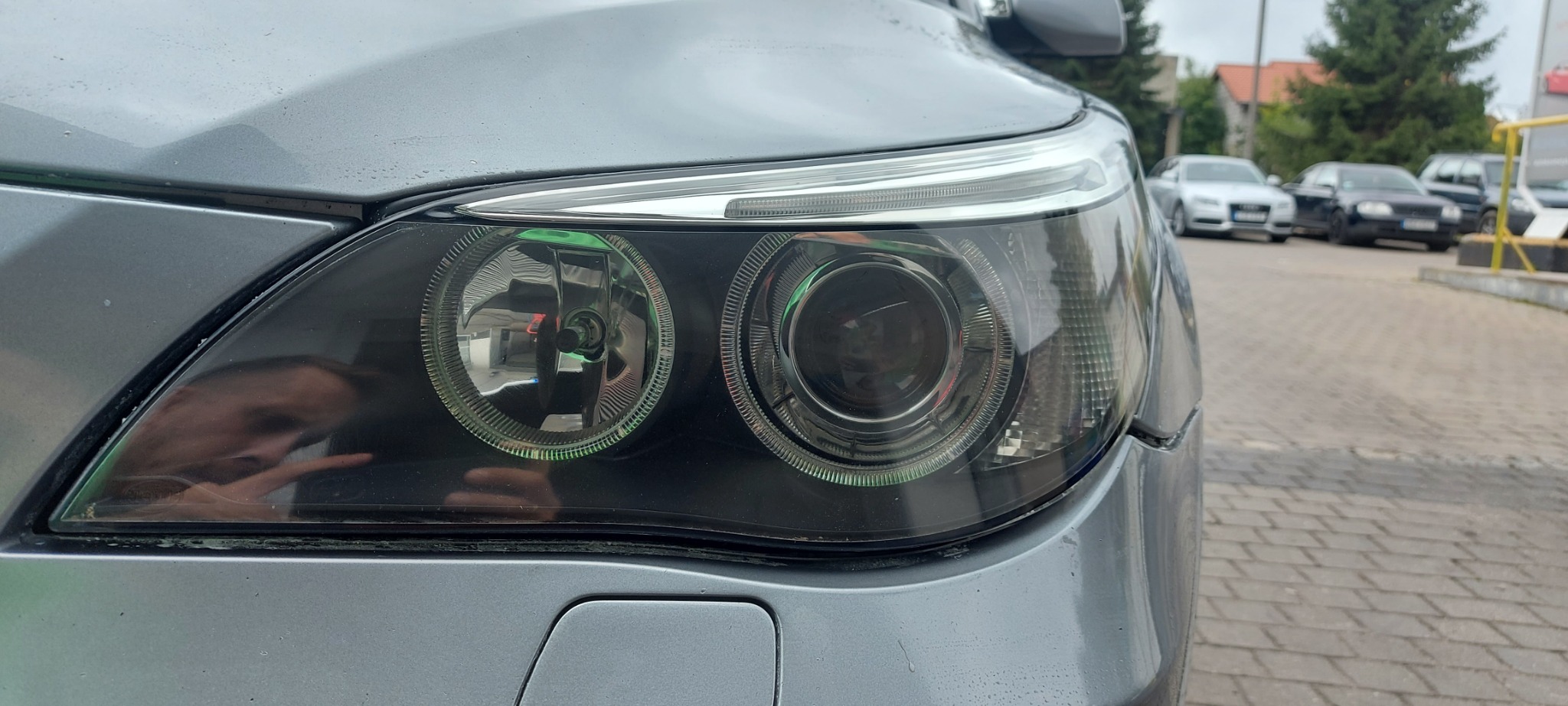 Szary samochód z widocznym przednim reflektorem, z odbiciem osoby w jego powierzchni.