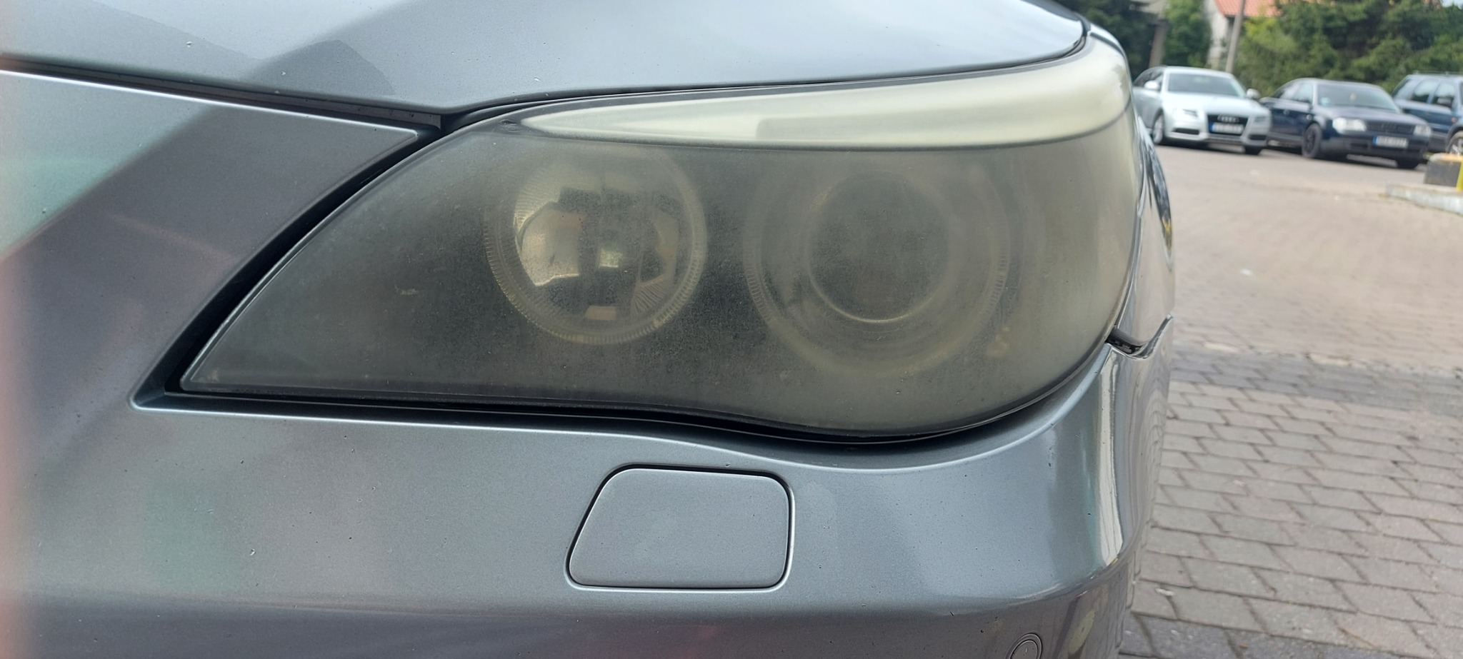 Silnie zmatowiałe reflektory przednie srebrnego samochodu osobowego, widoczne inne auta w tle.
