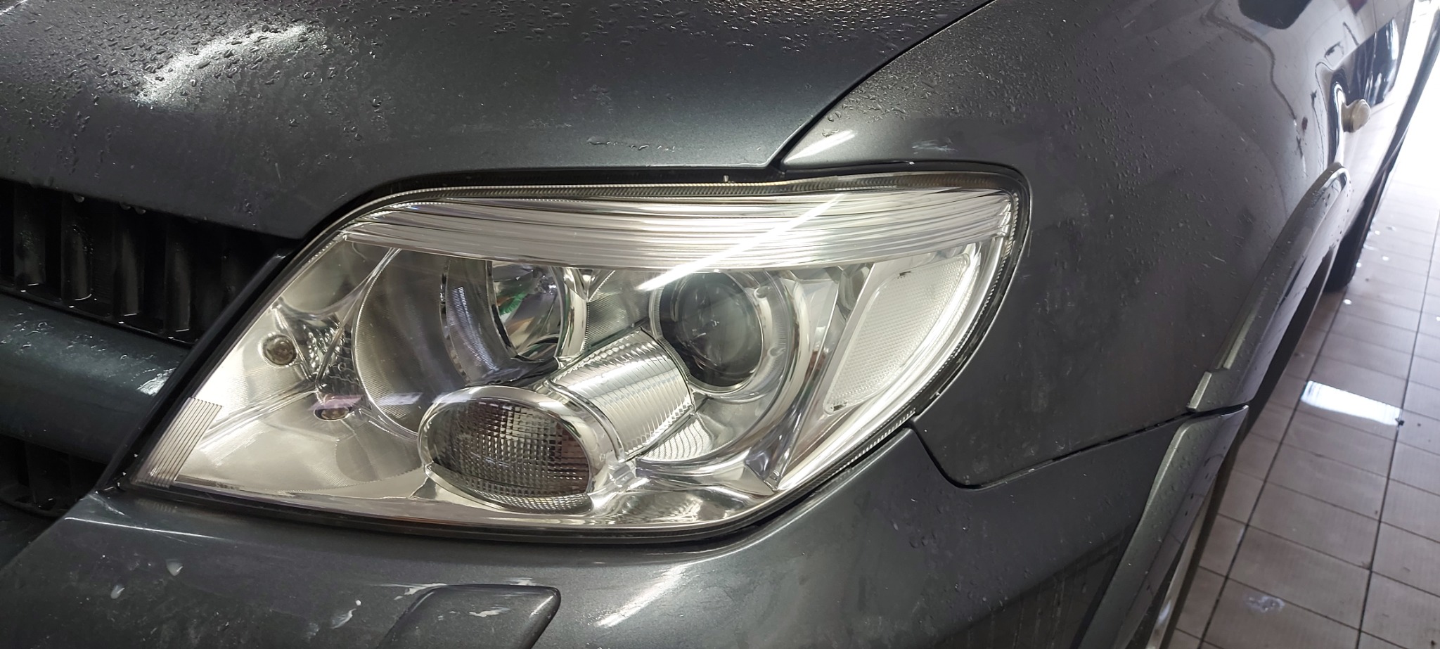 Błyszczący reflektor umytego, szarego samochodu z widocznymi kroplami wody na karoserii.