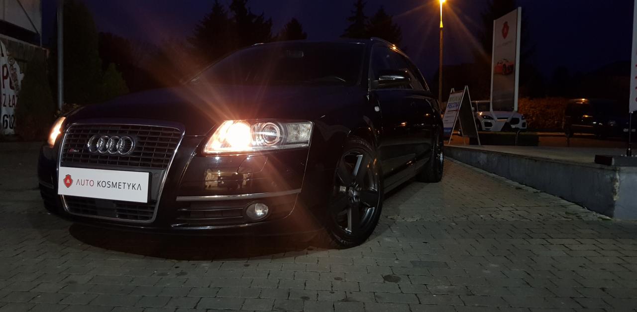 Ciemny samochód Audi kombi po usłudze auto kosmetyki, zaparkowany na brukowanym placu w nocy, z włączonymi światłami i logo firmy na tablicy rejestracyjnej.