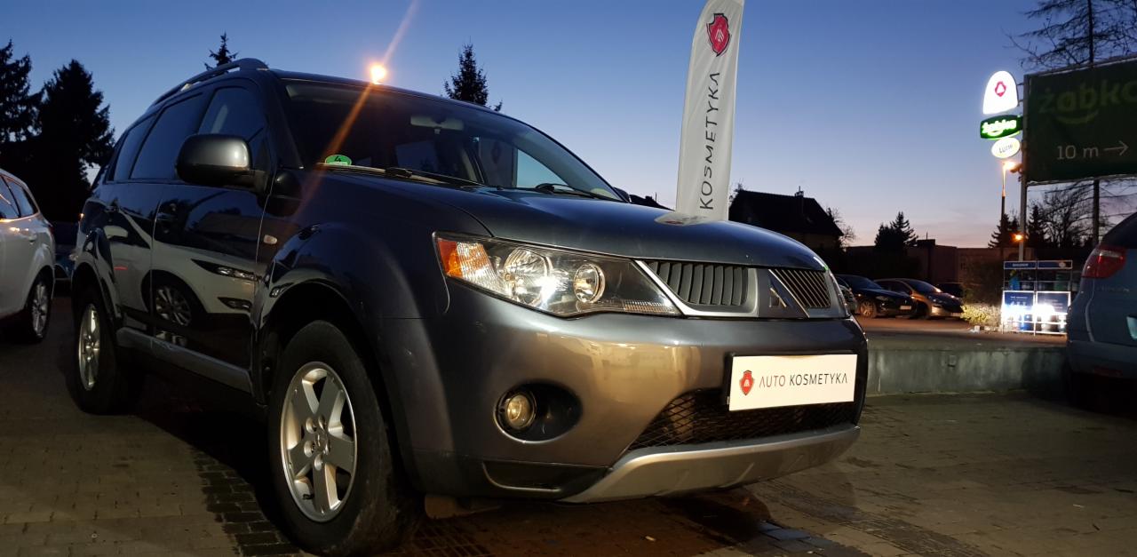Szary SUV Mitsubishi Outlander po usłudze auto kosmetyki, zaparkowany na zewnątrz z widocznym banerem reklamowym i logo firmy na przednim zderzaku, w tle sklep Żabka i inne samochody.