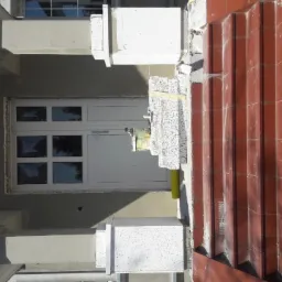 Wejście do budynku w trakcie prac wykończeniowych, widoczne płytki klinkierowe na schodach, styropianowe elementy dekoracyjne i białe drzwi wejściowe z szybami.