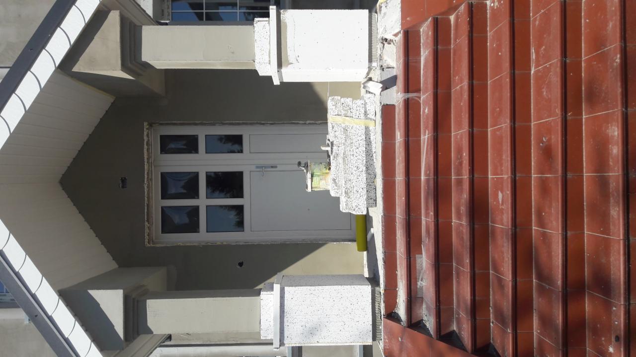 Wejście do budynku w trakcie prac wykończeniowych, widoczne płytki klinkierowe na schodach, styropianowe elementy dekoracyjne i białe drzwi wejściowe z szybami.