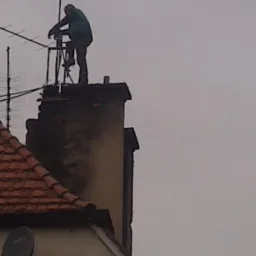 Mężczyzna na dachu, na drabinie, obok komina z antenami. Praca na wysokościach, serwis kominowy. Komin z widocznymi śladami użytkowania.