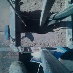 Widok z góry na rusztowanie z elementami komina w trakcie budowy, z fragmentem dachu z cegły i stopą pracownika w obuwiu roboczym.