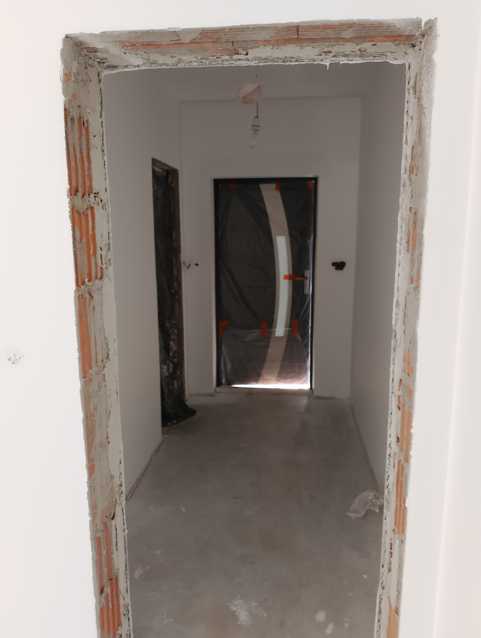 Surowy stan wykończenia wnętrza: widoczne cegły i resztki zaprawy wokół ościeżnicy, zabezpieczone folią drzwi w tle, goła żarówka zwisająca z sufitu.