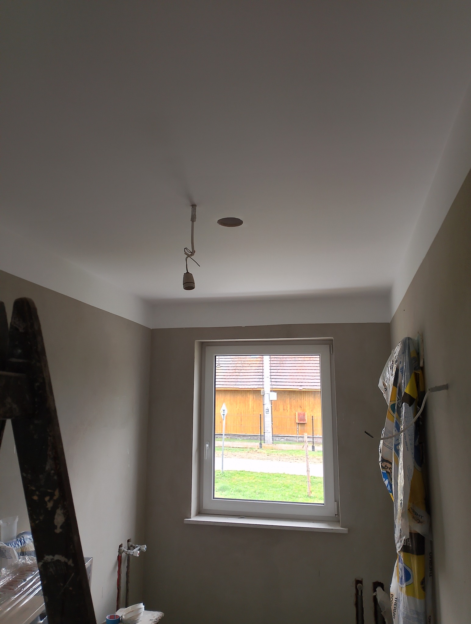 Pokój w trakcie prac wykończeniowych: świeżo pomalowany sufit z widocznym otworem na oświetlenie, ściany w kolorze beżowym, drabina oparta o ścianę po lewej stronie, okno z widokiem na podwórze...