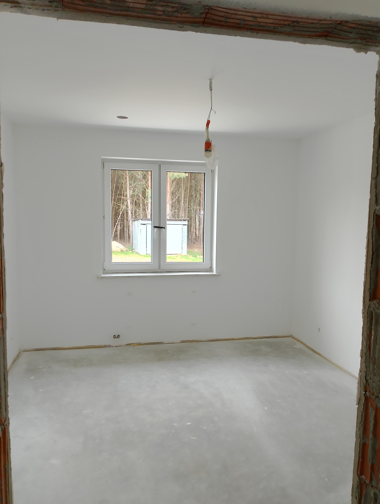 Pusty, jasny pokój z białymi ścianami i surową podłogą, widok przez surową, czerwoną ramę drzwi, okno z widokiem na drzewa na zewnątrz, przewody elektryczne zwisające z sufitu.
