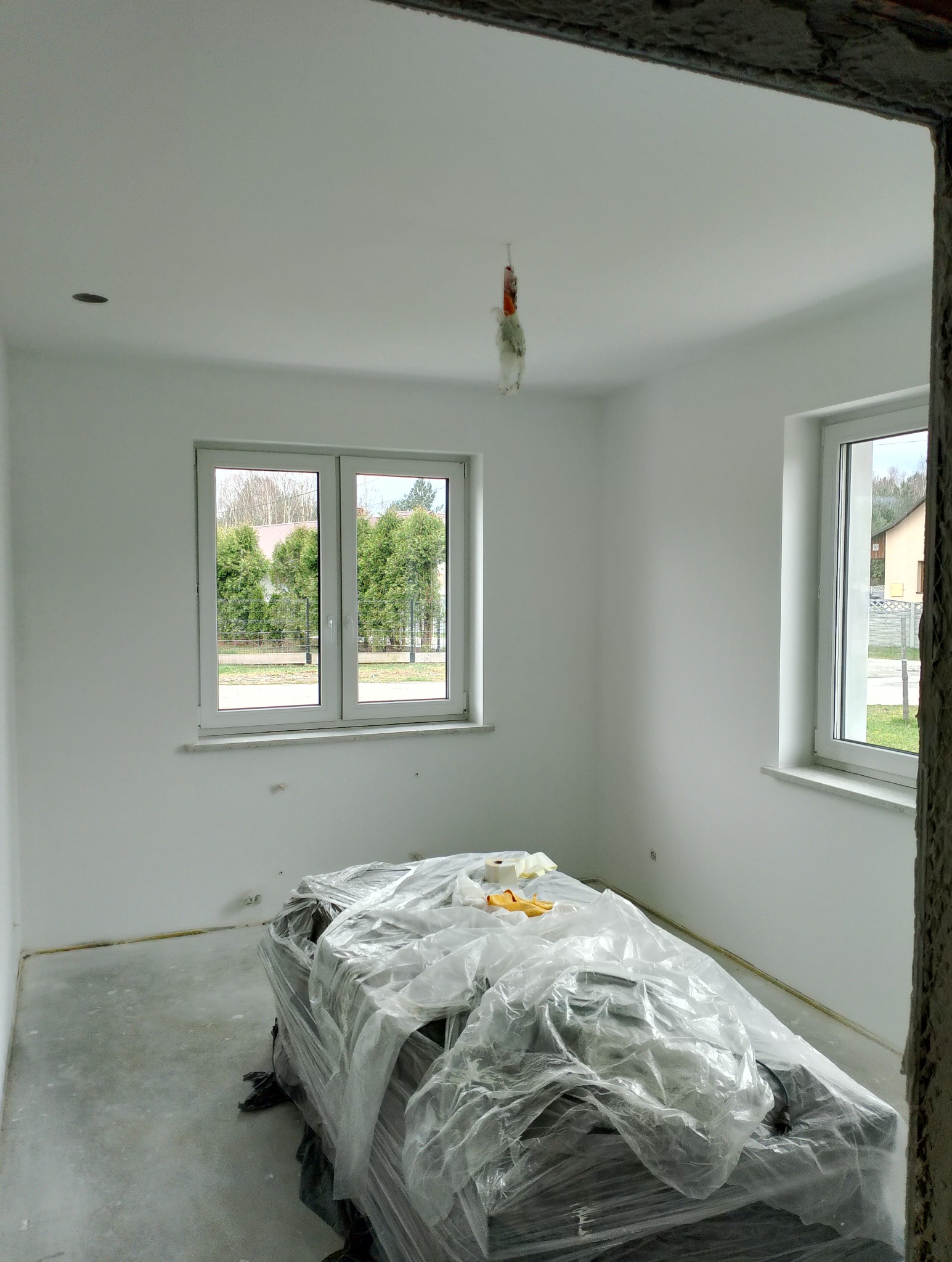 Pusta, świeżo wykończona na biało przestrzeń z dwoma oknami, z podłogą przygotowaną do dalszych prac i stertą materiałów budowlanych przykrytych folią na pierwszym planie, widoczne niedoskonałości...
