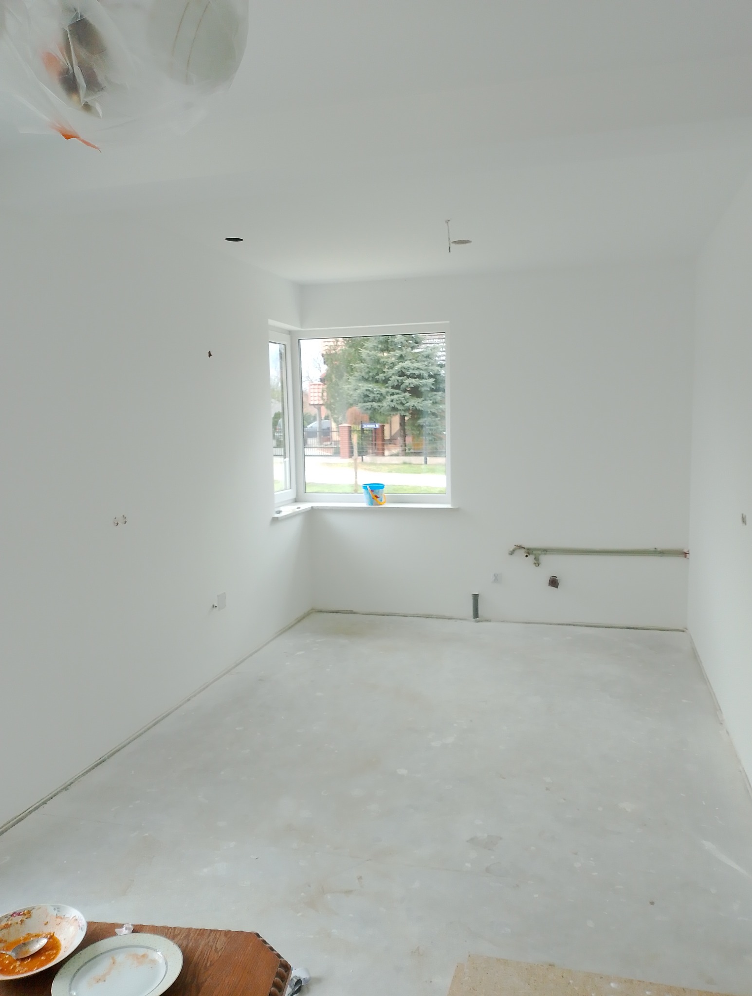 Pusty pokój z białymi ścianami po świeżym malowaniu, widoczne pozostałości po pracach na podłodze, okno z widokiem na zieleń, niebieskie wiaderko na parapecie, wystające rury.