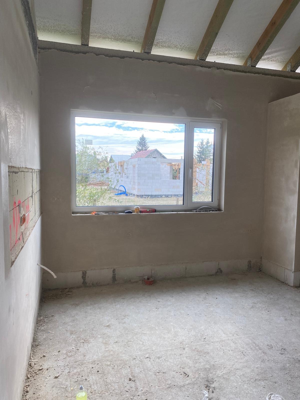Pokój w trakcie prac wykończeniowych: ściany pokryte świeżym tynkiem, widoczne narzędzia na parapecie okna, surowy beton na podłodze, w tle budowa domu w stanie surowym.