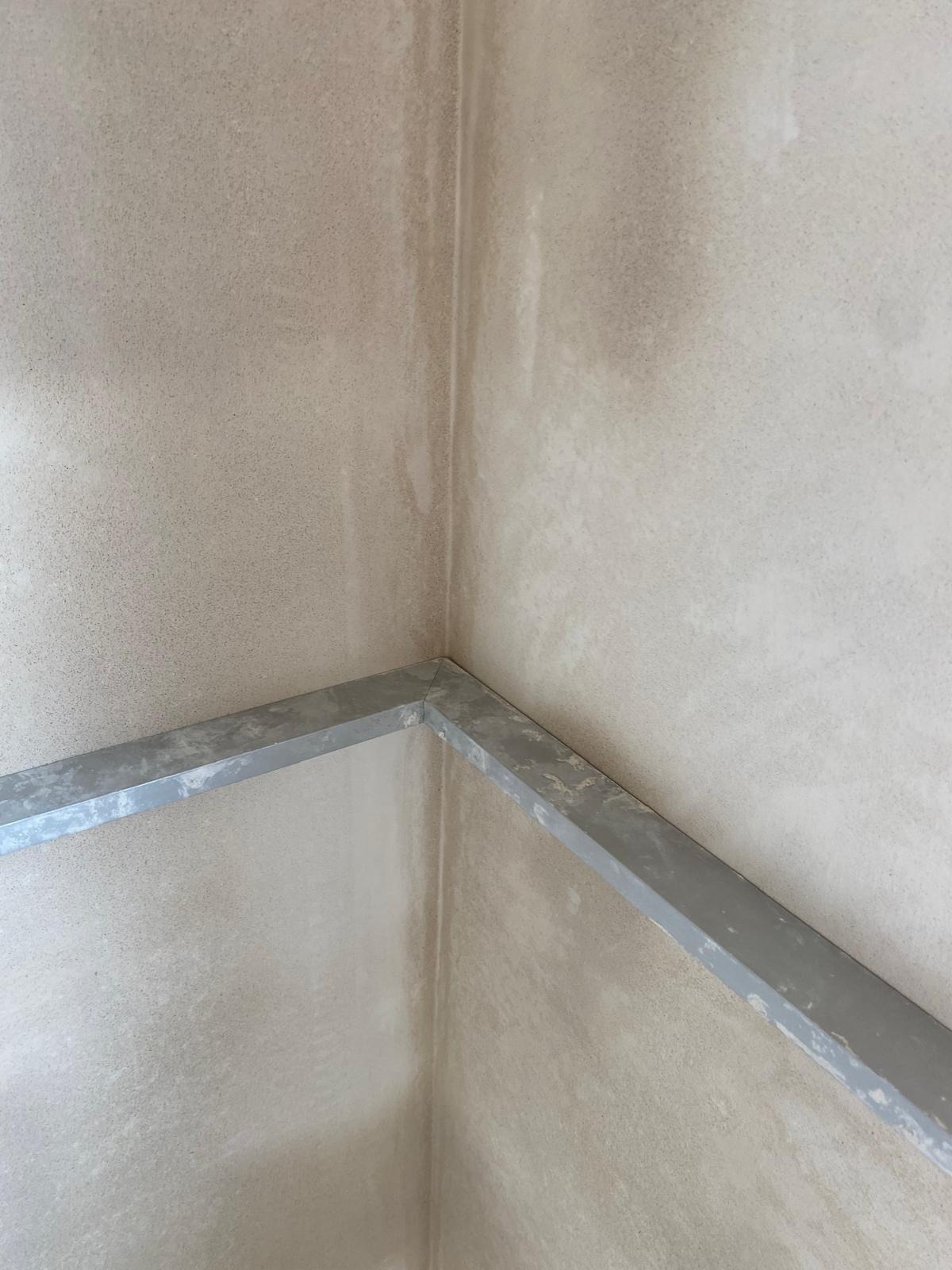 Fragment ściany w rogu pomieszczenia po nałożeniu warstwy szpachli, widoczny metalowy profil narożnikowy.