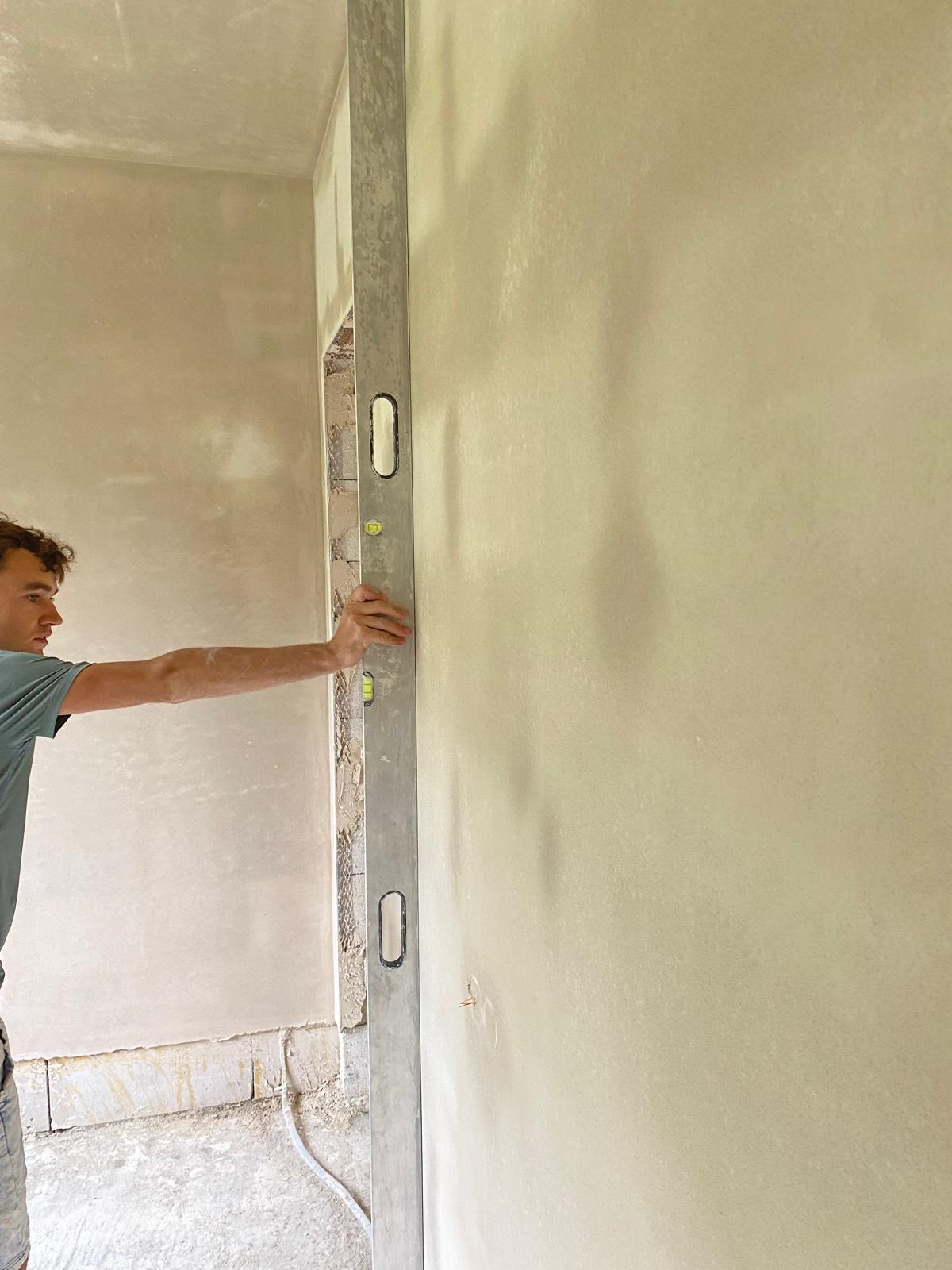 Mężczyzna przykłada poziomicę do świeżo wyszpachlowanej ściany, widoczne ślady po szpachlowaniu i niedokończone wykończenie przy podłodze.