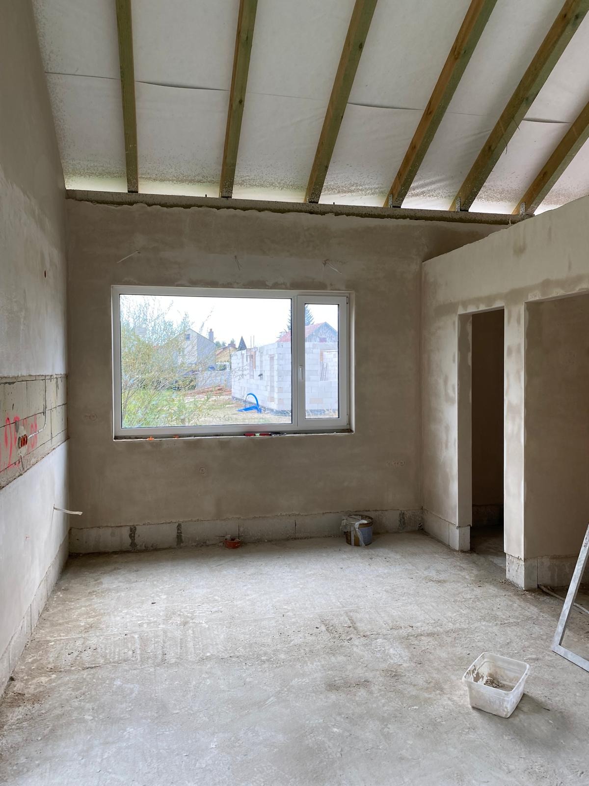 Surowe wnętrze w trakcie prac: ściany po wstępnym szpachlowaniu, odsłonięta konstrukcja dachu z drewnianymi belkami, okno z widokiem na budowę. Na podłodze resztki materiałów budowlanych...