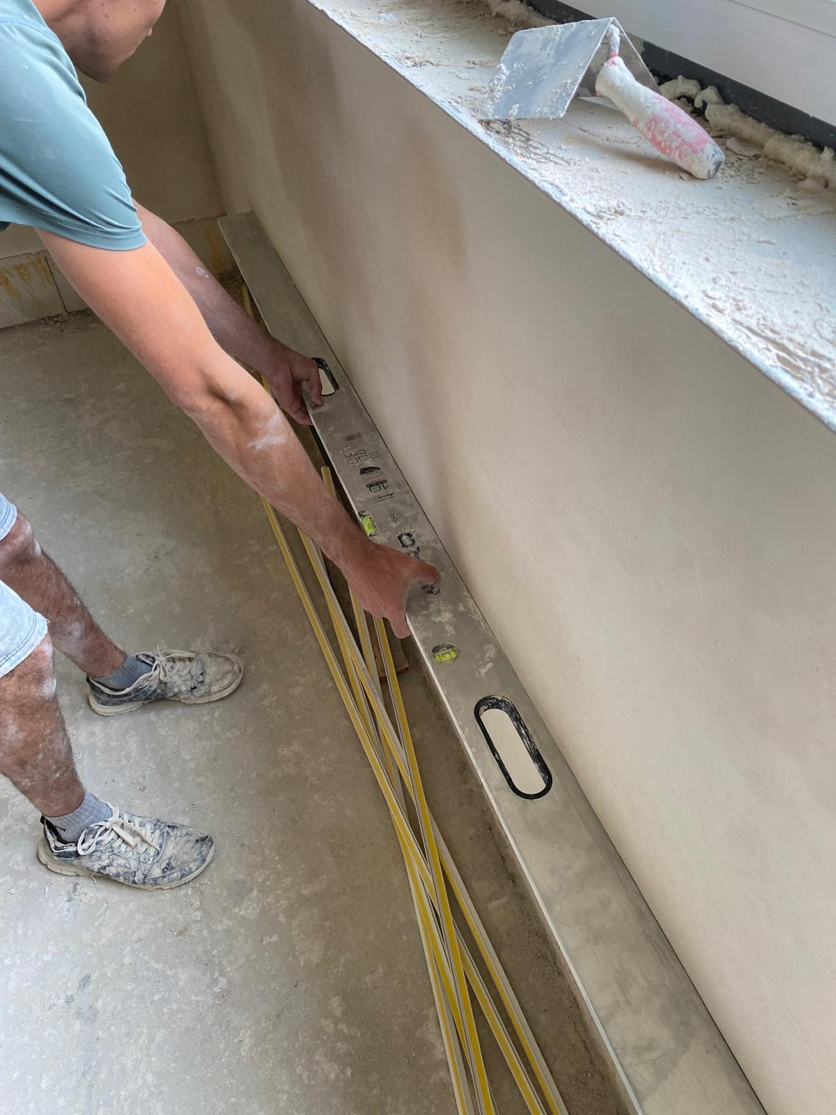 Pracownik przykłada długą poziomicę do świeżo wyszpachlowanej ściany, obok leżą żółte listwy przypodłogowe, na parapecie brudna szpachelka.