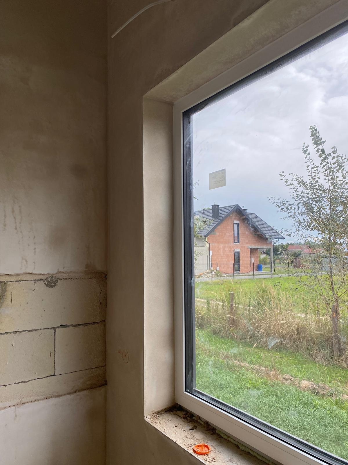 Surowa ściana z bloków i świeżo nałożona gładź szpachlowa przy oknie z widokiem na budynek w stanie surowym otwartym i zarośnięte pole.