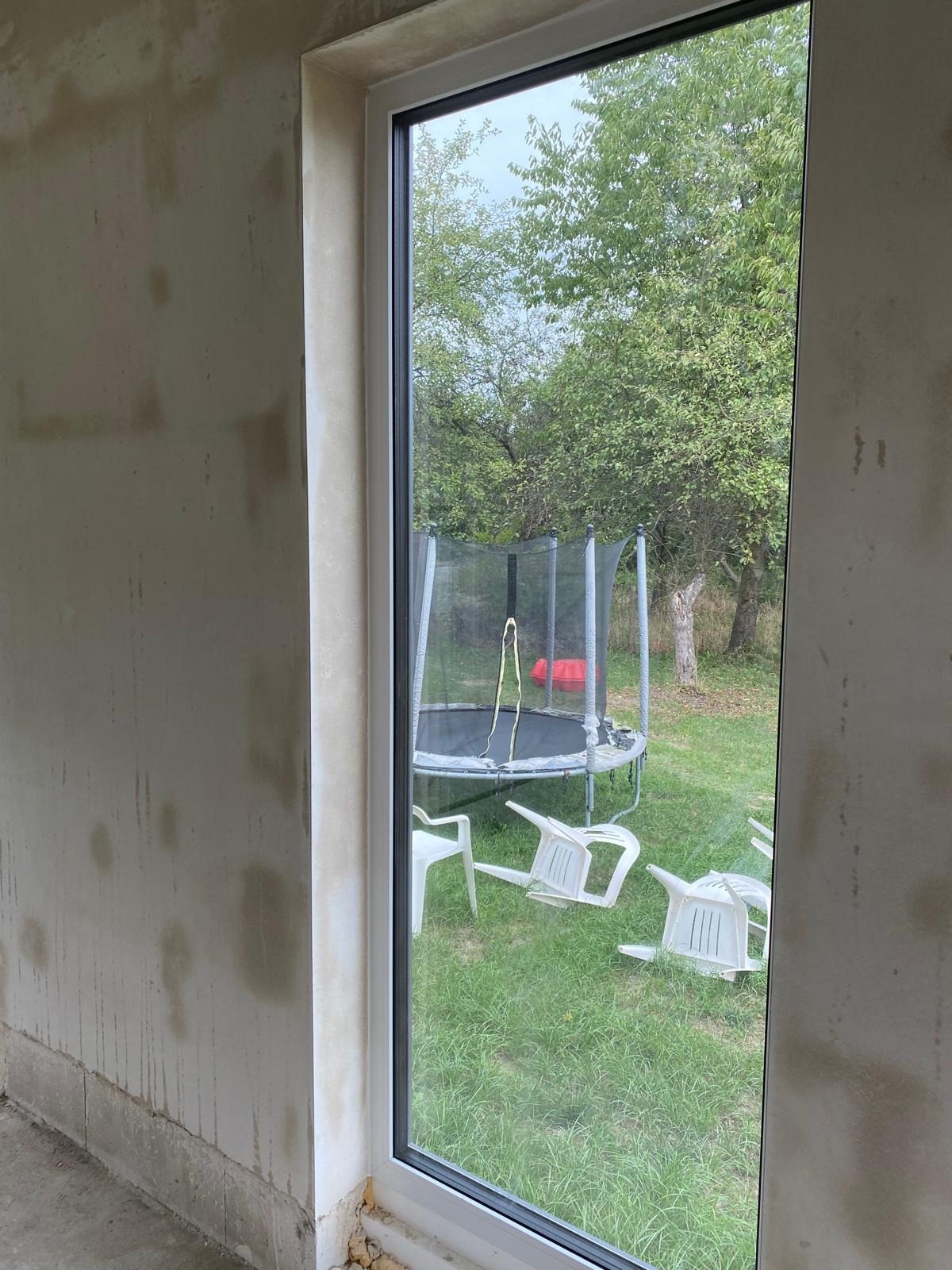 Ściana wewnętrzna w trakcie prac przygotowawczych, widoczne ślady po wcześniejszych warstwach i naprawach, okno z widokiem na ogród z trampoliną i porozrzucanymi plastikowymi krzesłami.