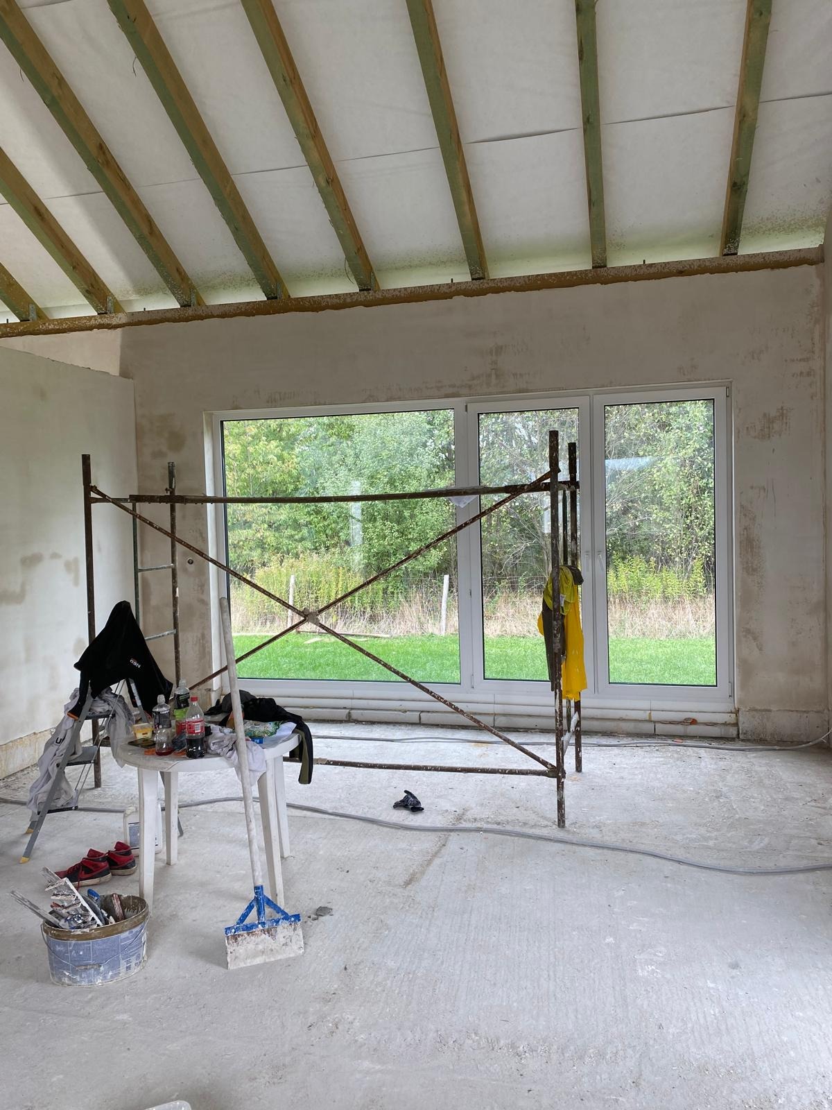Wnętrze w trakcie prac wykończeniowych: ściany po nałożeniu gładzi, widoczne rusztowanie, okno z widokiem na zieleń, narzędzia i akcesoria malarskie na stoliku, odsłonięta konstrukcja dachu.