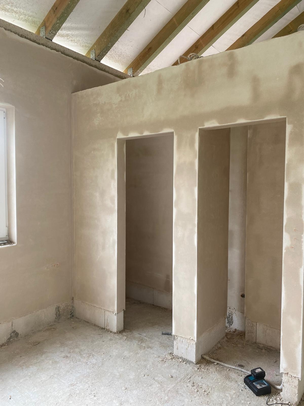 Wnętrze w trakcie prac przygotowawczych: surowe ściany po nałożeniu szpachli, widoczne dwa otwory drzwiowe, niezagruntowany sufit z drewnianą konstrukcją, na podłodze resztki materiałów i narzędzia.